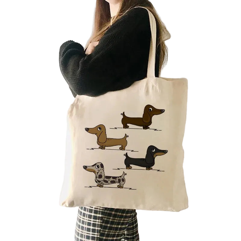 Person carrying a cream canvas tote bag featuring four illustrated Dachshund dogs in different coat colours